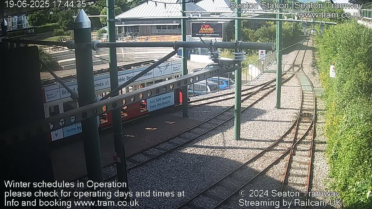 A tram is parked at a station with train tracks branching off, surrounded by greenery under sunny conditions.