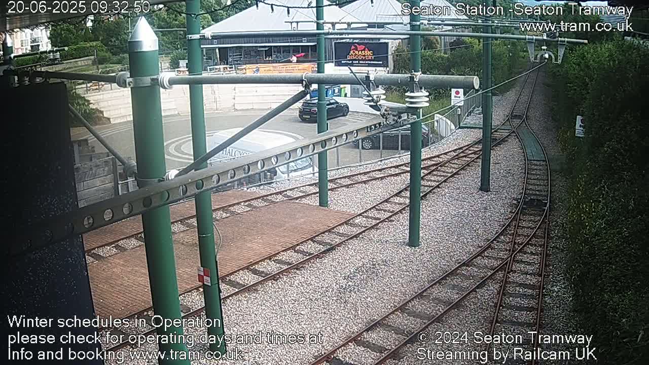 A tram station with several cars parked nearby, featuring a curving tram track and a gravel surface, under a partly cloudy sky.