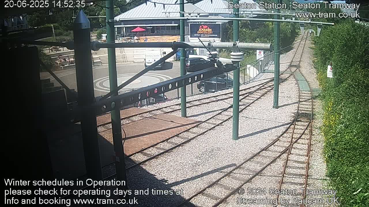 A sunny overhead view of a tram station with train tracks curving away from the camera, several parked cars, and a building in the background.
