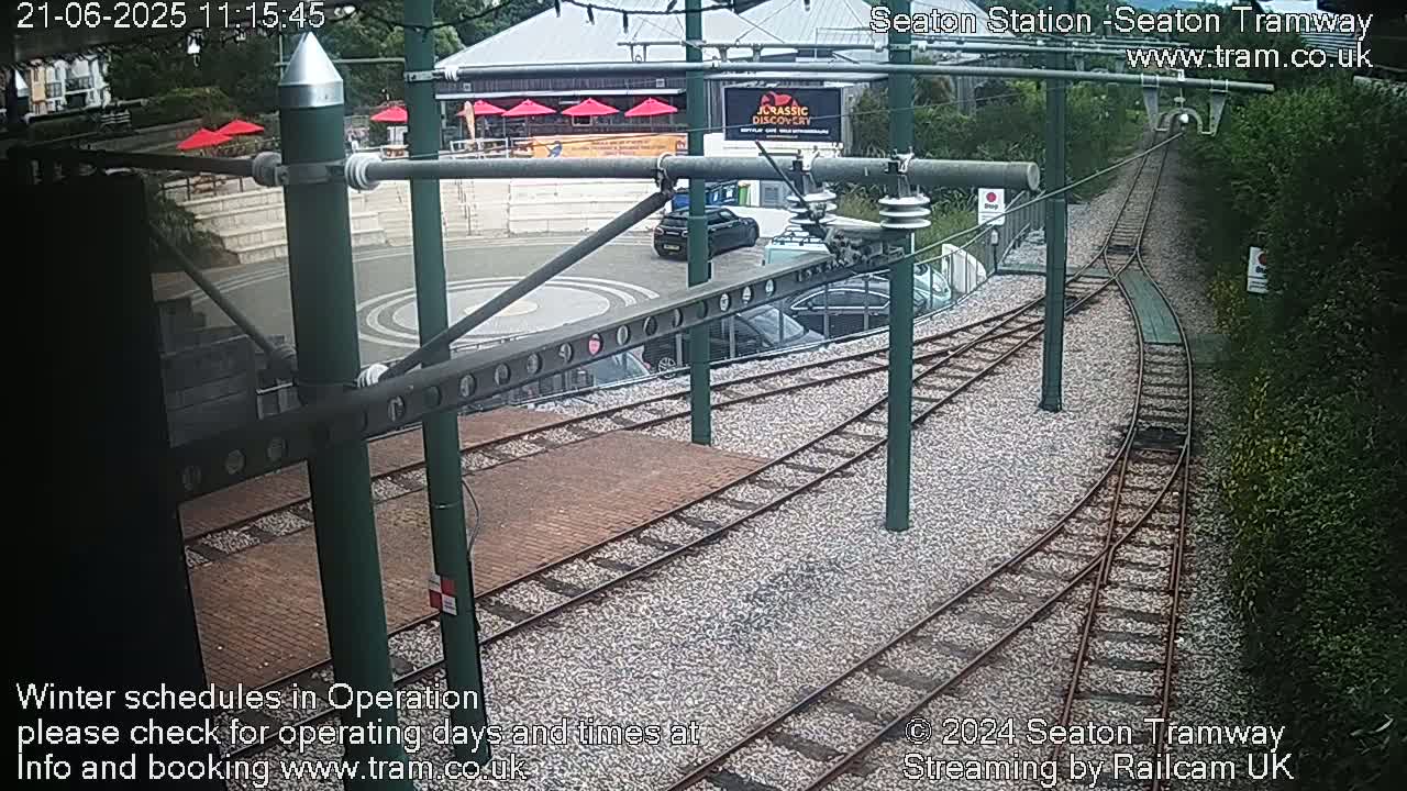 A high-angle view shows a tram station with train tracks curving through a gravel area, some parked cars, and a building with red umbrellas in the background under overcast skies.