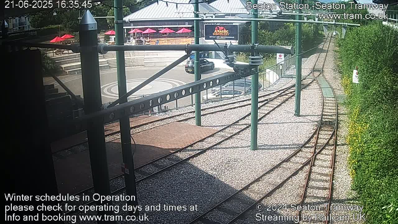 A sunny outdoor view of a tram station with converging tracks, a nearby building, and parked cars.