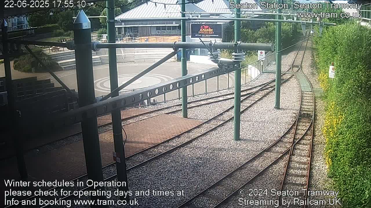 Tram tracks curve around a paved area with seating, beside a building and a green hedge, in sunny weather.