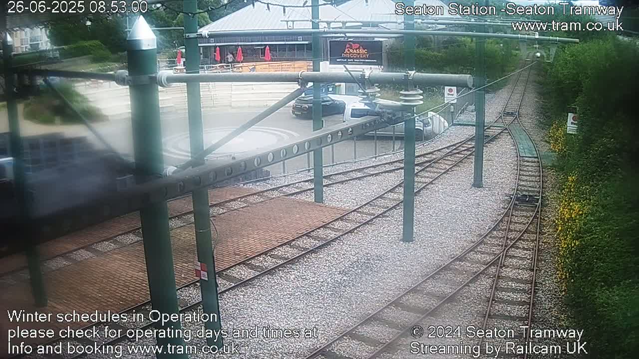 Tram tracks curve through a station area with a building and some cars visible, bordered by greenery under overcast skies.