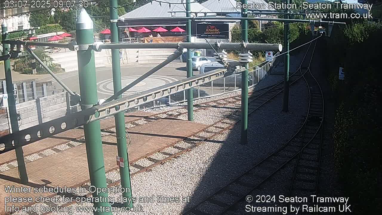 A sunny outdoor scene shows tram tracks curving around a paved area with some buildings and cars visible, next to a wooded area.