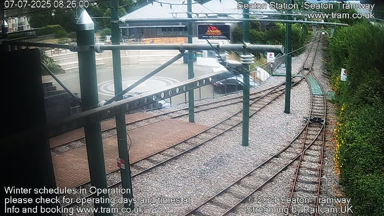 Tram tracks curve through a station area next to a building and greenery, under a mostly clear sky.