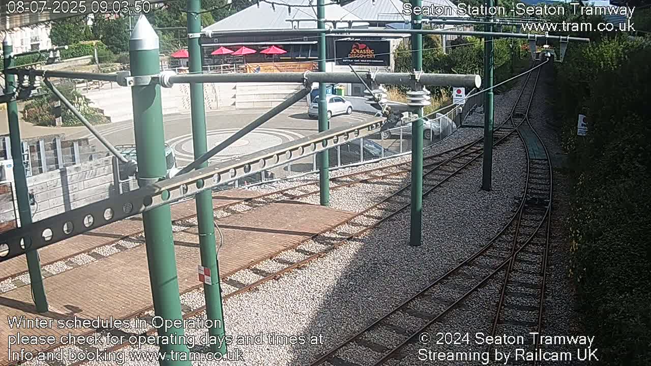 A sunny view of a tram station with train tracks and a parking lot, showing a partly visible building and surrounding vegetation.