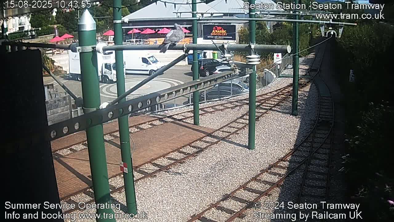 A high-angle view of a tram station with several sets of tracks curving away from a paved area, with some cars parked nearby, under overcast skies.