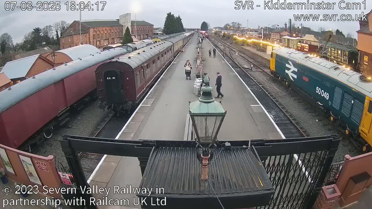 A train station platform with several passenger cars and a diesel locomotive under an overcast sky.