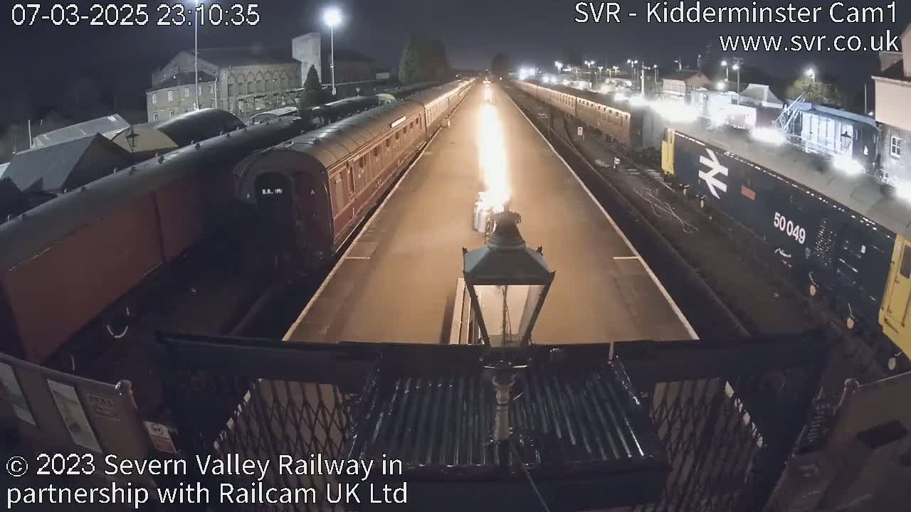 At night, a long train sits at a station platform next to another train, with a bright flame emanating from a source between them.