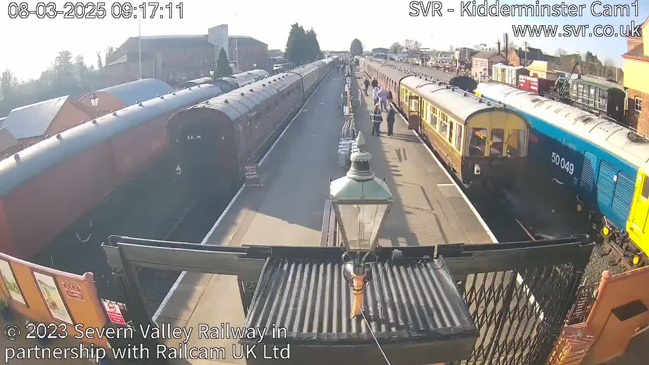 On a sunny day, several passenger train cars are stopped at a station platform with a few people standing nearby.