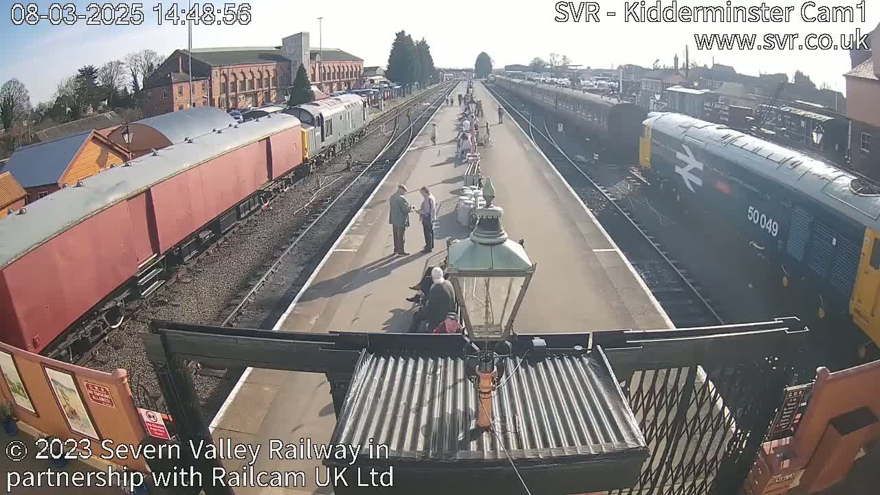A sunny day at a train station shows several people standing on a platform between two sets of train cars, one set of which is maroon.