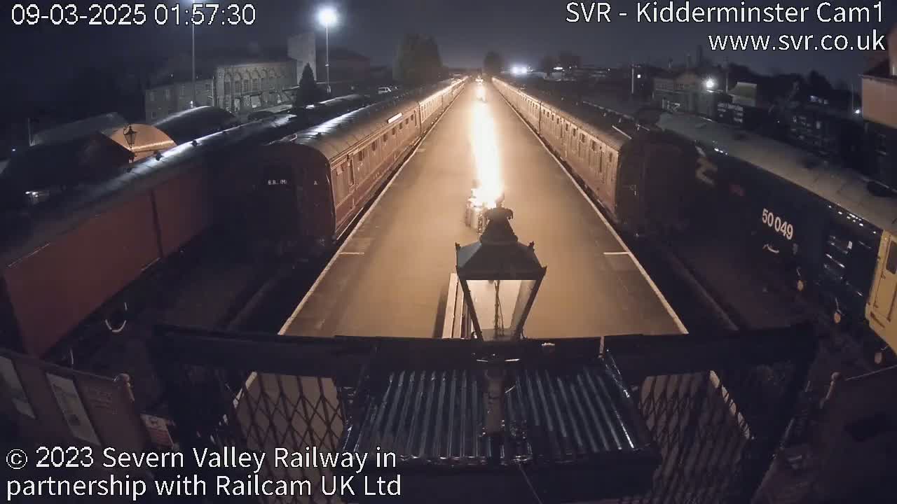 At night, two long lines of train cars are parked facing each other on a platform with a bright flame between them.