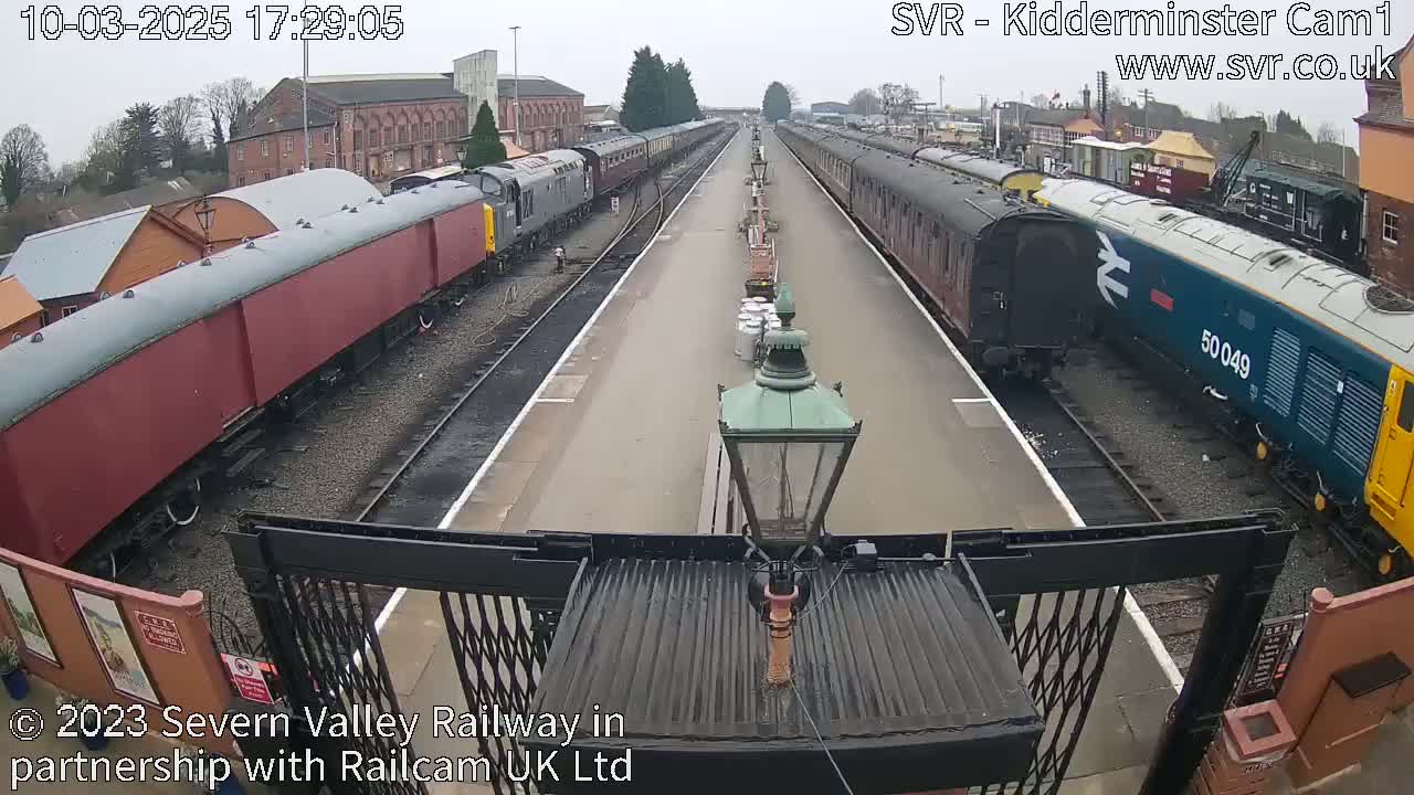 A high-angle view of a railway station platform on an overcast day with multiple train cars, some red and some blue, parked on either side of the tracks.