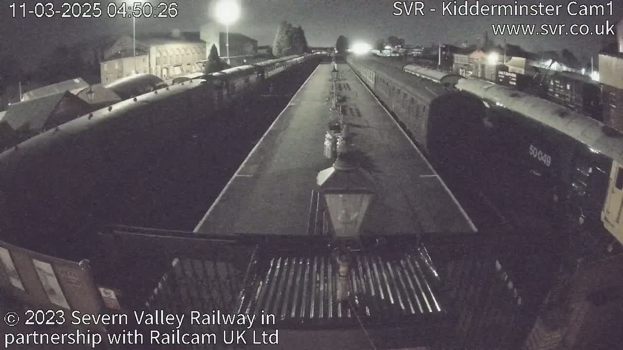 A nighttime view of a train station platform with multiple train cars parked along the tracks under dim lighting.