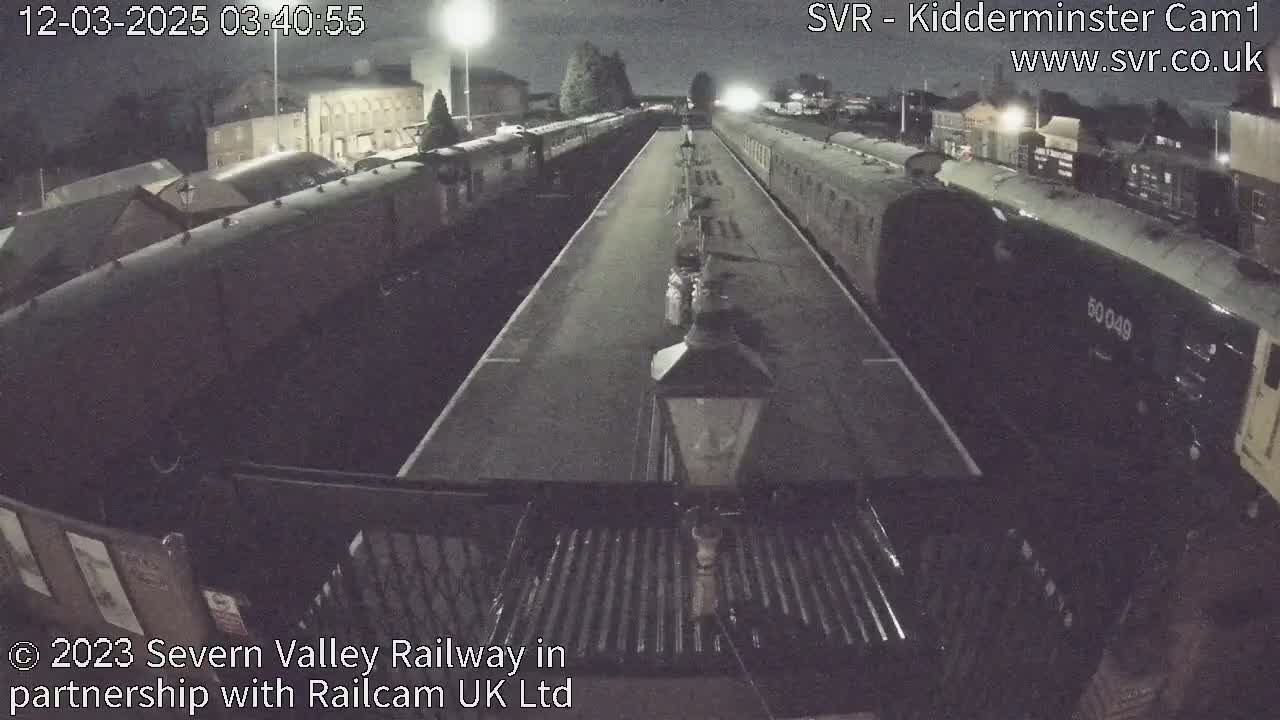 A nighttime, overhead view shows a railway station with several passenger trains parked on either side of a long platform under artificial lighting.