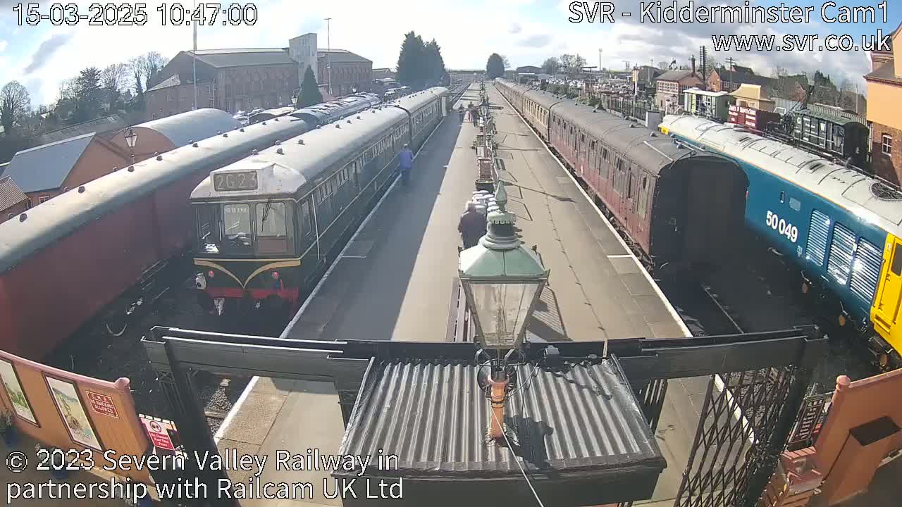 A sunny day at a railway station shows several vintage passenger train cars and a modern diesel locomotive parked on parallel tracks.