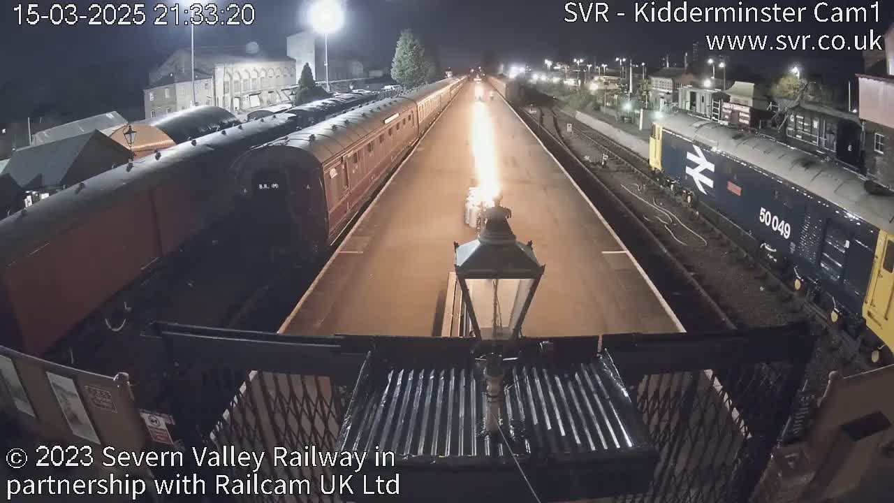 At night, a long train is parked at a station platform next to a diesel locomotive, with a bright flame visible in the middle of the tracks.