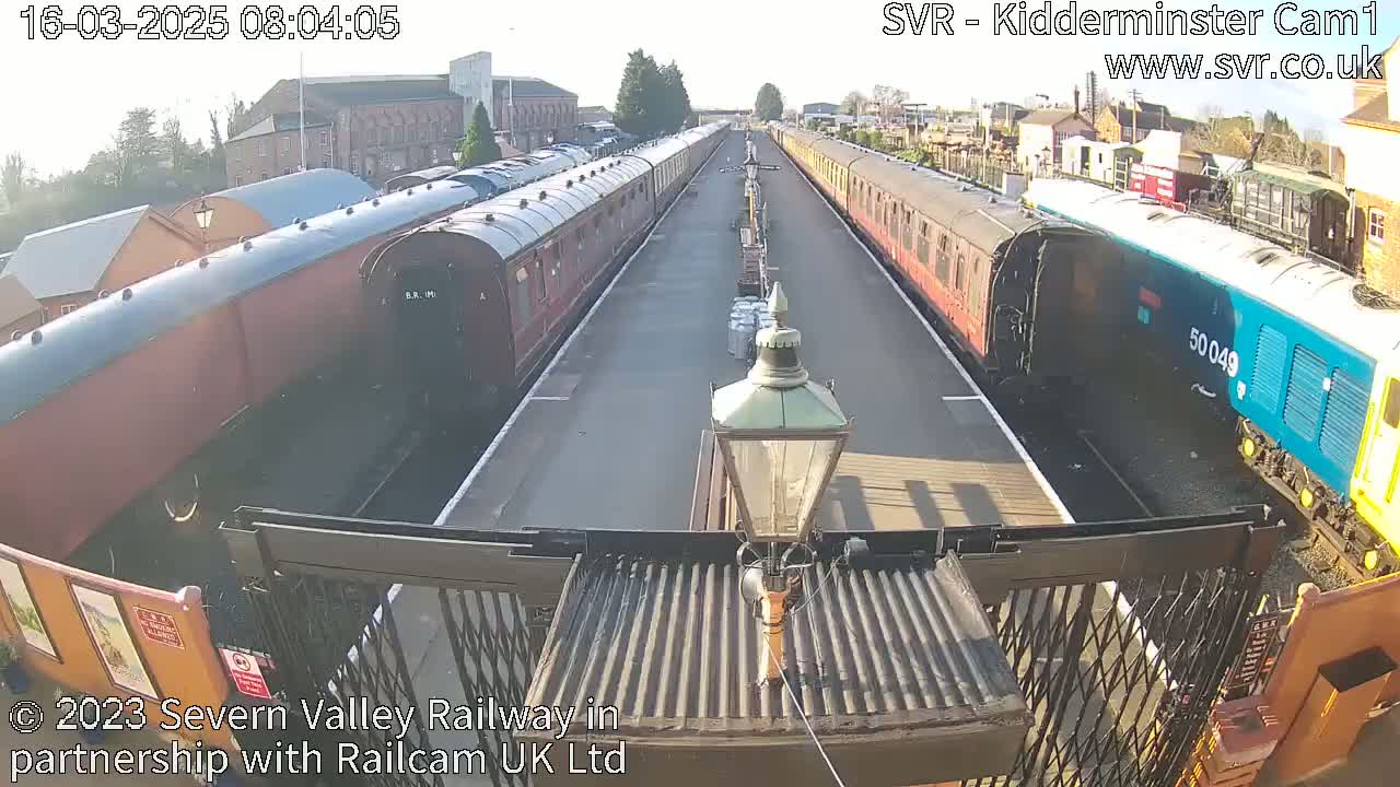 A sunny day at a train station shows two lines of vintage railway carriages facing each other, with a modern diesel locomotive at the end of one line.
