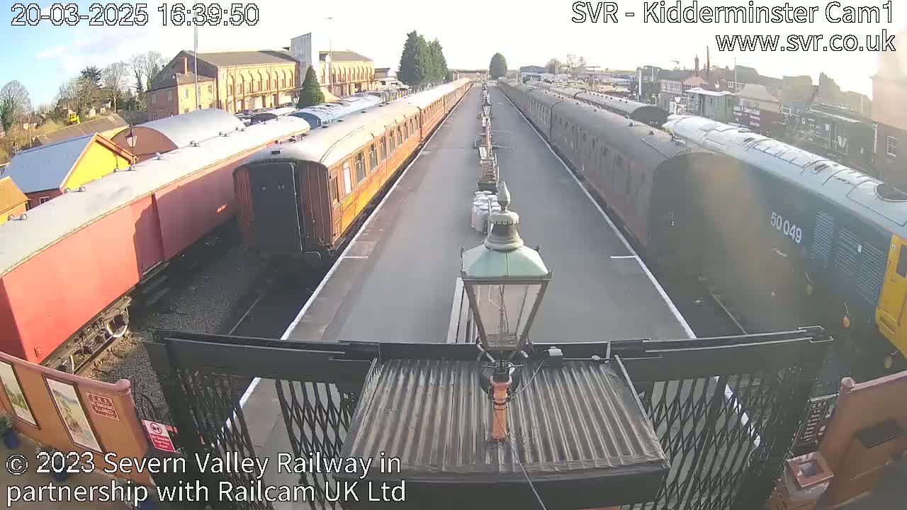 Two long passenger trains are parked on parallel tracks at a railway station on a sunny day.