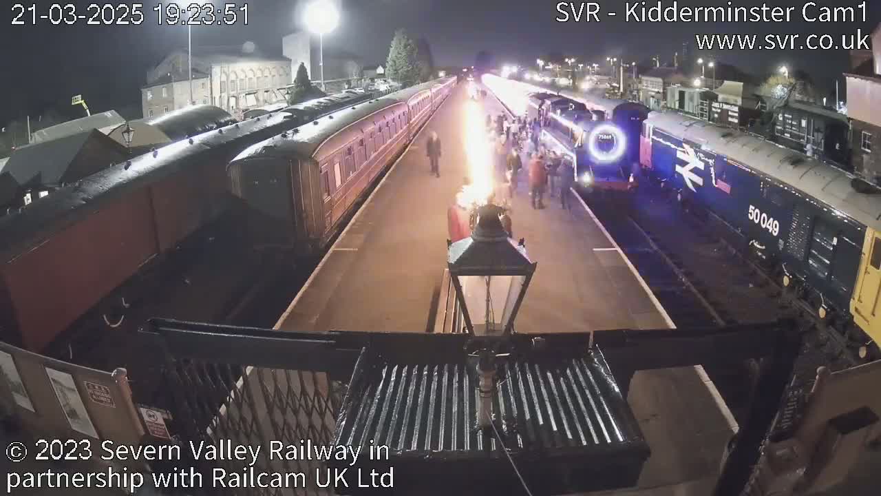 At night, a large group of people stand between two trains on a railway platform, a flame burning between them.
