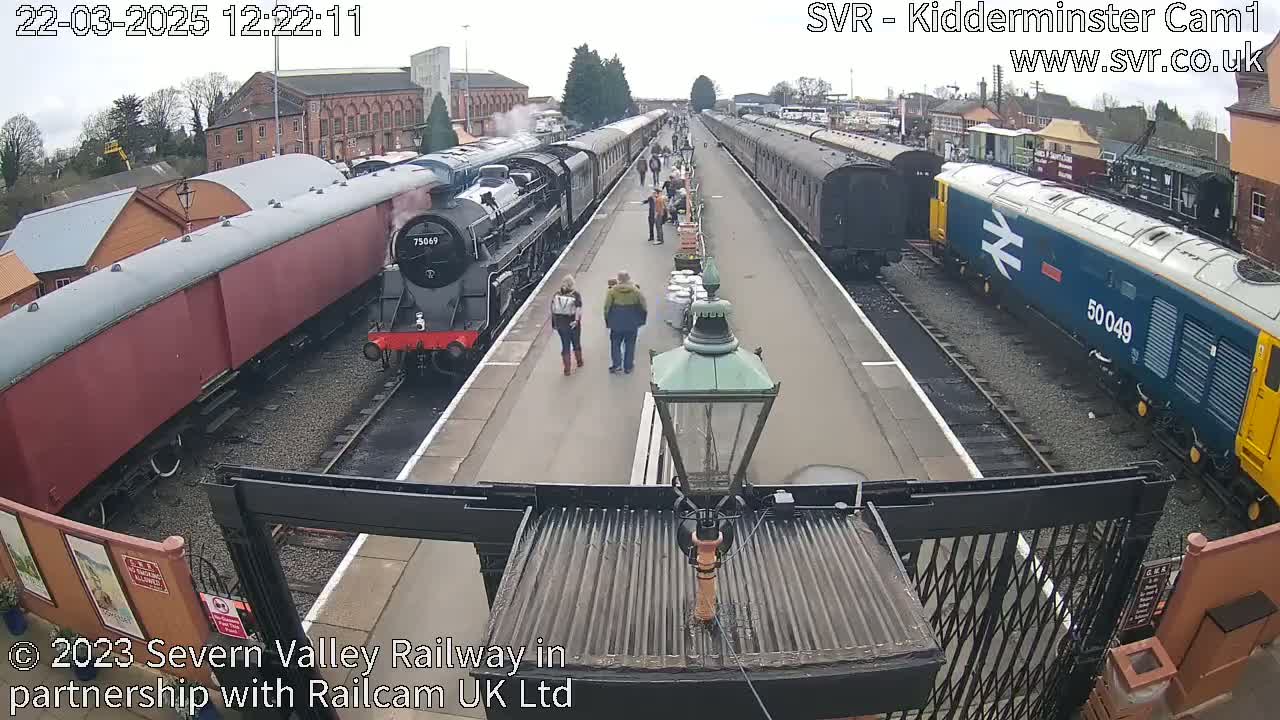 A steam train and a diesel train are at a station platform with several people walking around on a partly cloudy day.