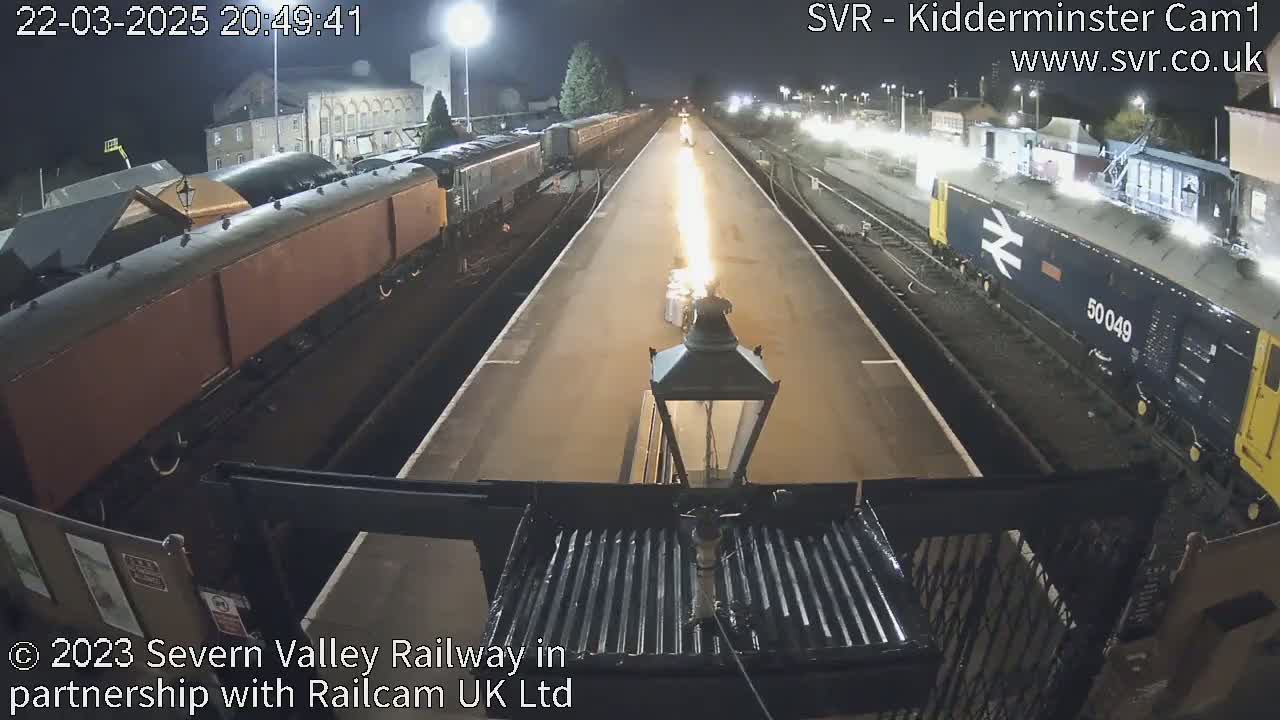 At night, a train platform shows two trains and a line of fire down the center, illuminated by overhead lights.