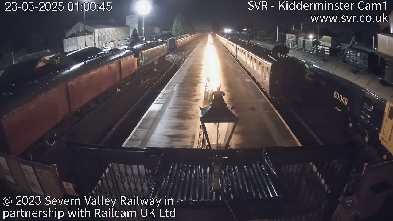 Two trains, one dark-colored and one reddish-brown, are parked at a station at night, with a bright light emanating from between them illuminating a wet platform.