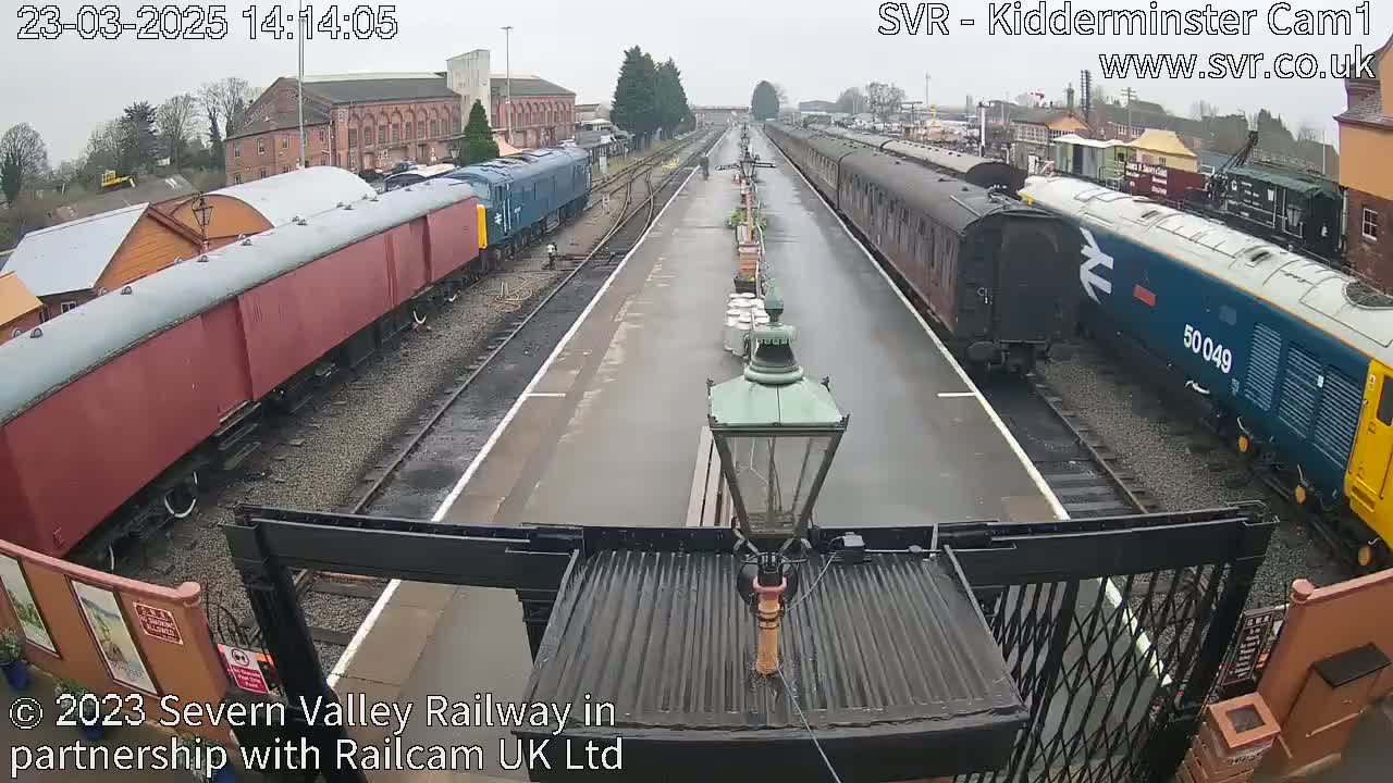 Two long trains, one red and one blue and yellow, are parked on rain-slicked railway tracks in front of older buildings.