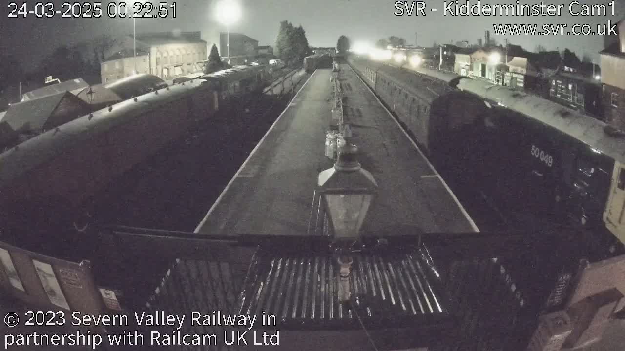 A nighttime view of a train station platform with several train cars parked alongside it under a dark sky.