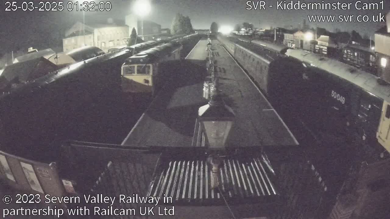 A nighttime view of a railway station platform with several trains parked along the tracks under dark, possibly overcast skies.