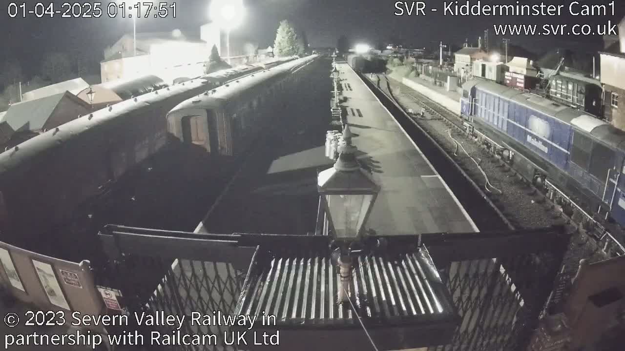 At night, a train station platform shows several passenger rail cars and a diesel locomotive under bright artificial lighting.