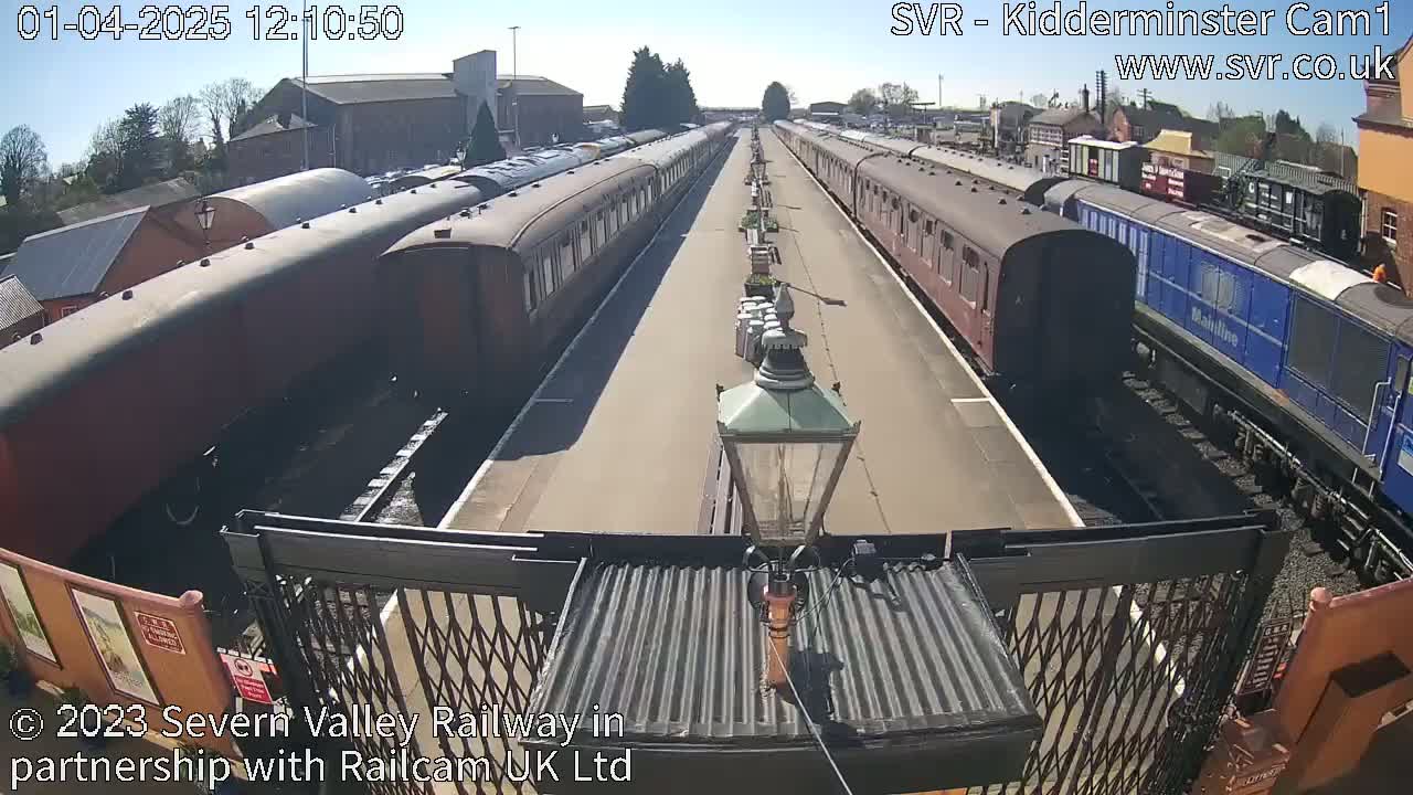 Two long lines of passenger train cars are parked on parallel tracks on a sunny day, with a blue diesel locomotive at the end of one line.