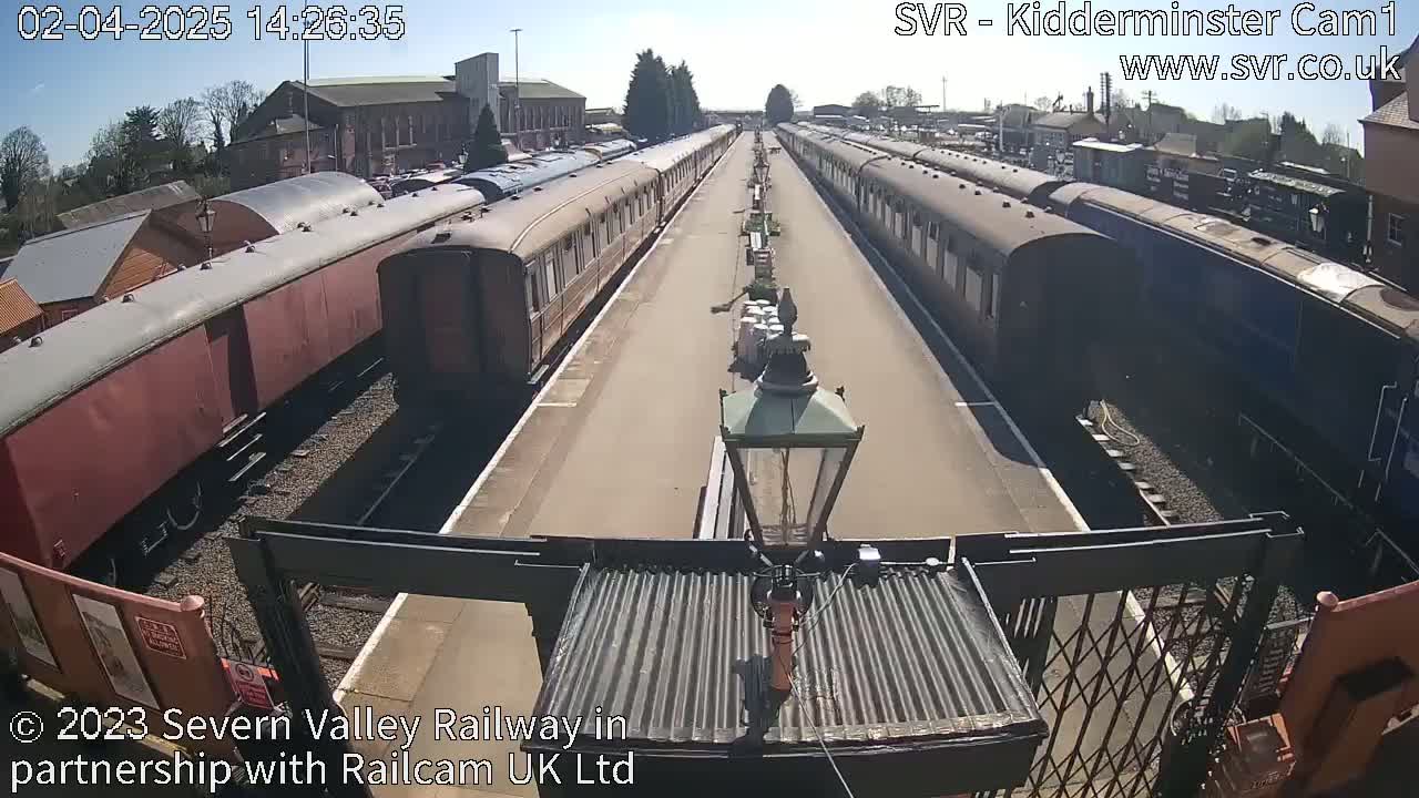 Under a sunny sky, two long lines of passenger train cars are parked on parallel tracks at a railway station.
