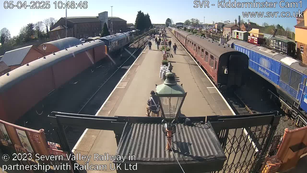 A sunny day at a train station shows multiple passenger cars and locomotives, with people walking along the platform.