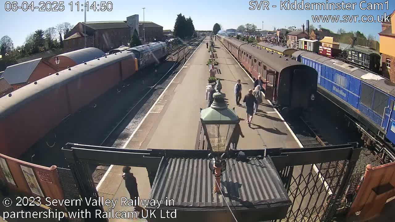 A sunny day at a train station shows several people walking on the platform between two lines of railway cars.