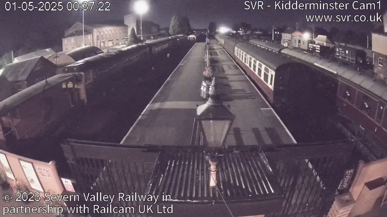 At night, two long trains are parked at a station platform, with buildings visible in the background under a dark sky.