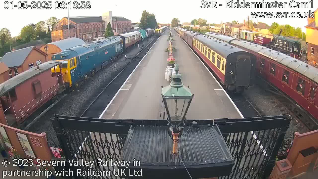 A railway station platform with multiple passenger train cars and a diesel locomotive parked on either side, under a partly cloudy sky.