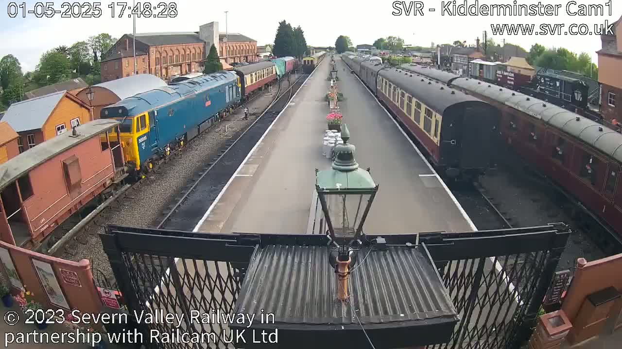 A high-angle view of a train station on a partly sunny day shows several railway carriages and locomotives on parallel tracks.