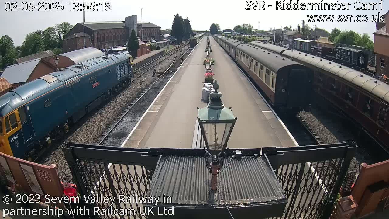 A blue diesel locomotive and a long line of passenger cars are parked at a train station on a sunny day.