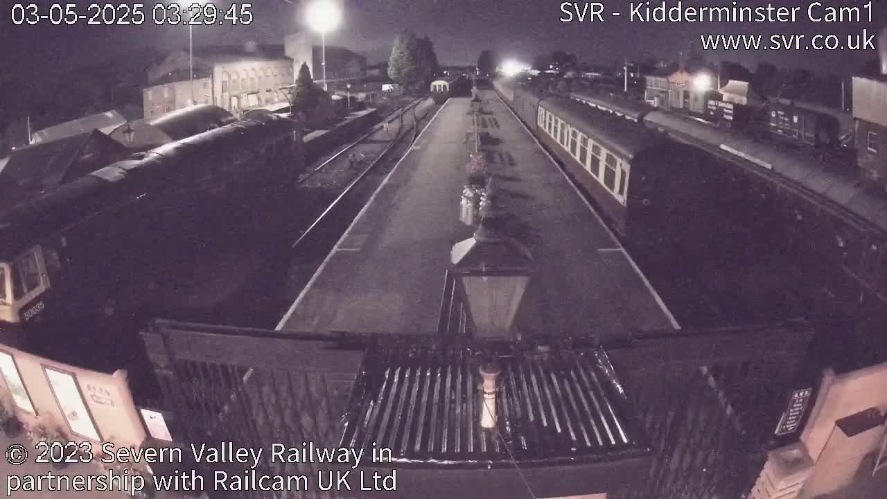 At night, a railway station platform shows two trains, one diesel and one passenger, parked along the tracks.