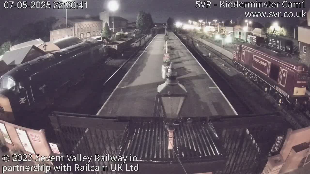 At night, two locomotives are visible at a train station platform, with one dark-colored locomotive on the left and a maroon-colored locomotive on the right.