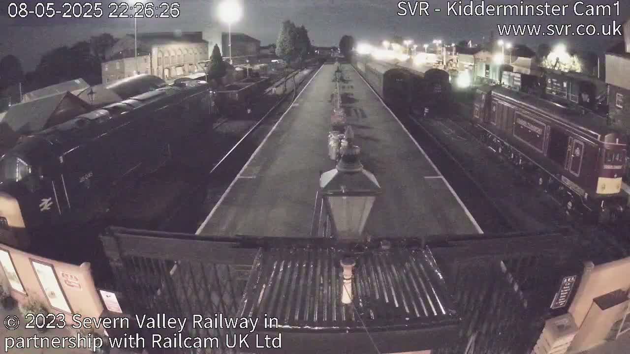 At night, two dark-colored locomotives are parked on train tracks at a station, with a brightly lit platform separating them.