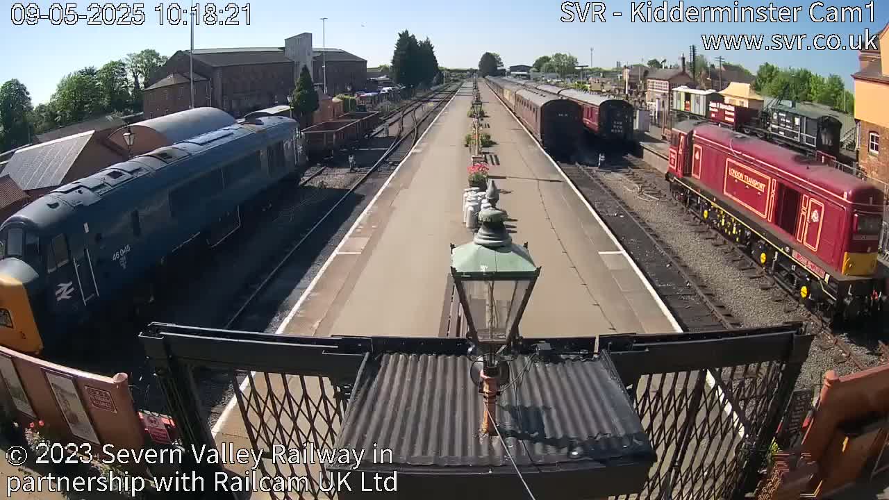 A sunny day at a train station shows two diesel locomotives and several railway carriages on parallel tracks.