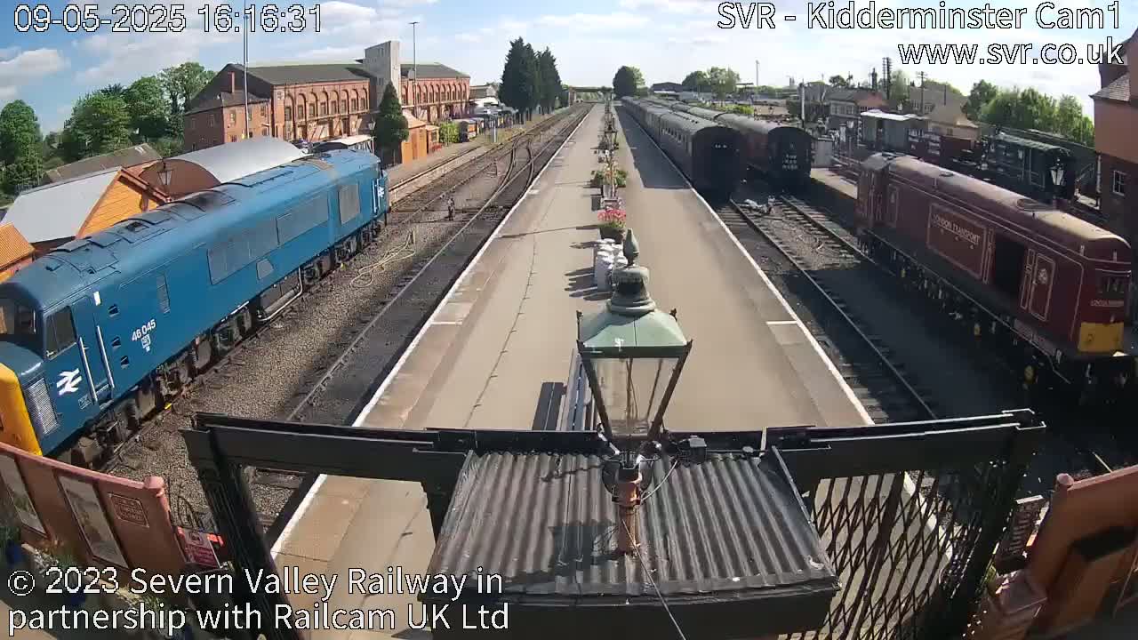 A sunny day at a railway station shows a blue diesel locomotive and a maroon diesel locomotive, with several railway carriages and brick buildings visible in the background.