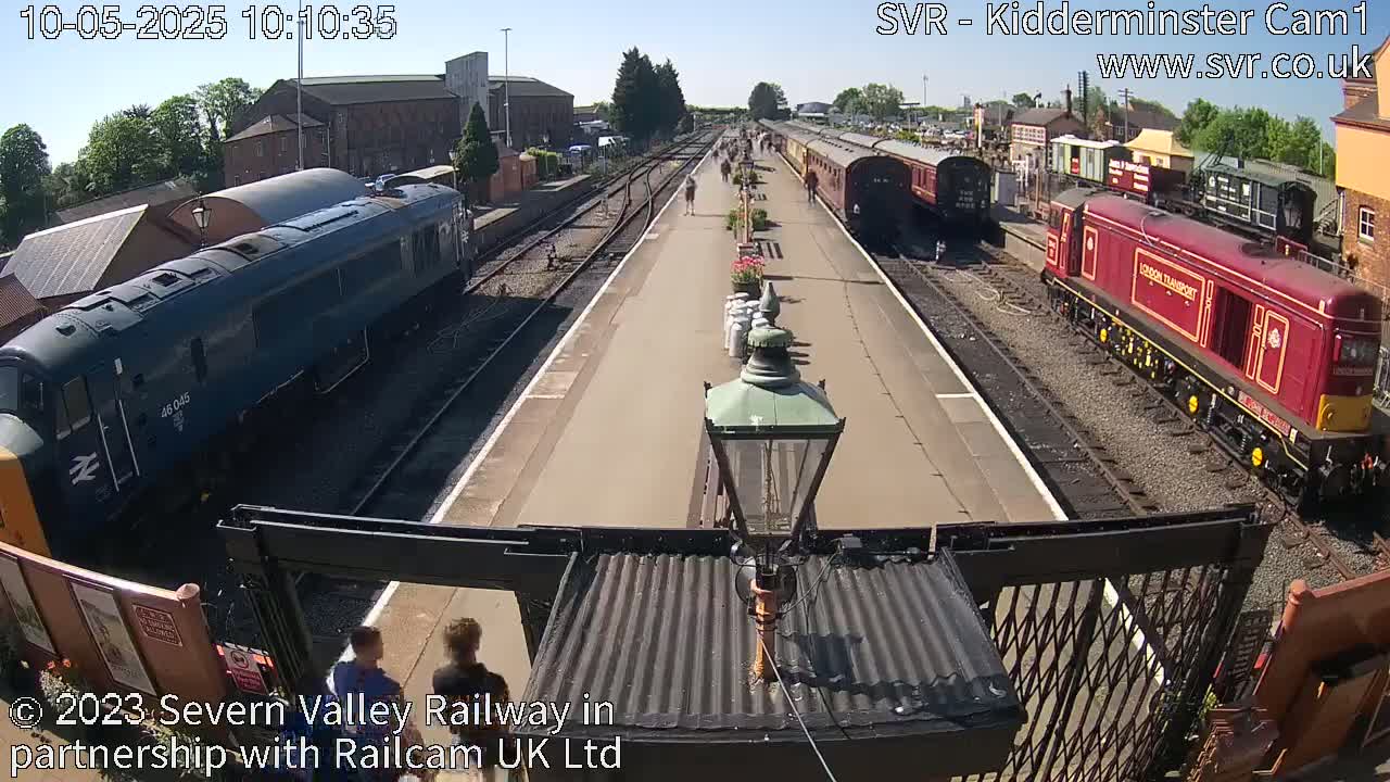 A sunny day at a train station shows two locomotives and several passenger cars on the tracks, with people walking on the platform.