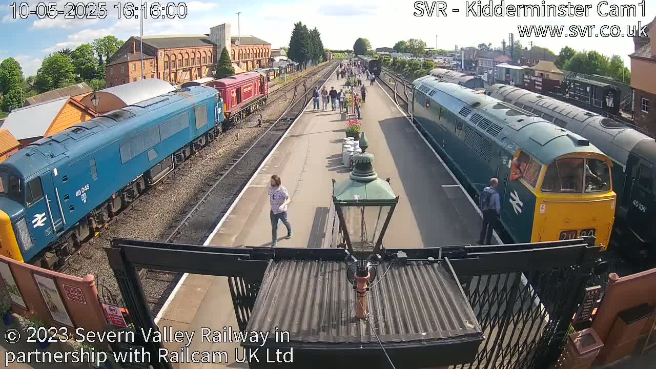 On a sunny day, multiple diesel locomotives are parked at a train station platform where people are walking around.