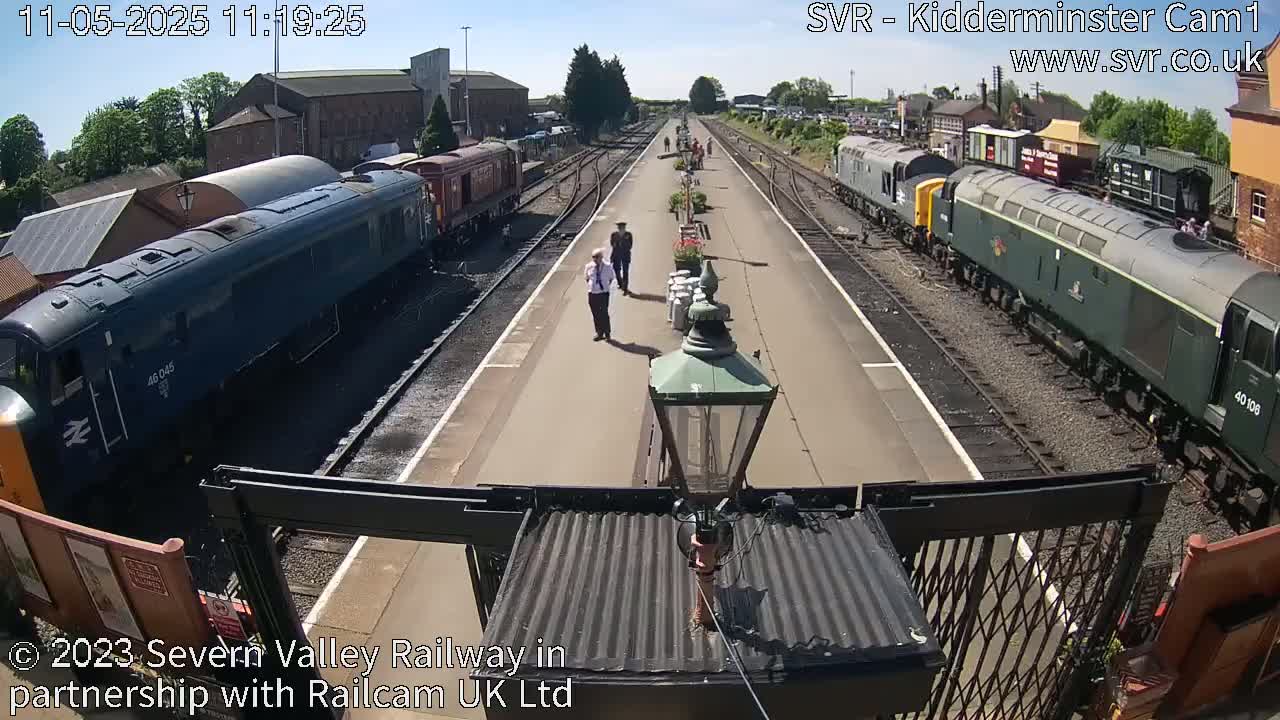A sunny day at a train station shows several locomotives on parallel tracks, with a few people standing on the platform between them.