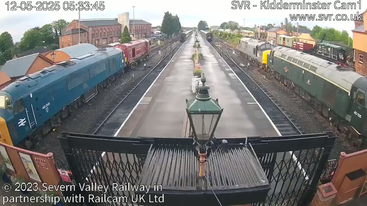 A wet railway station platform with several diesel locomotives parked on either side of the tracks.