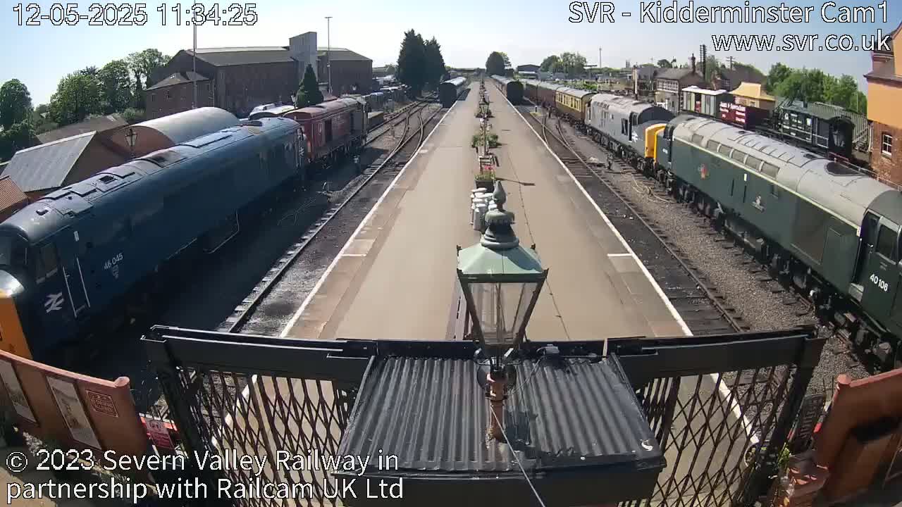 A sunny day at a train station shows multiple locomotives and passenger cars on parallel tracks.