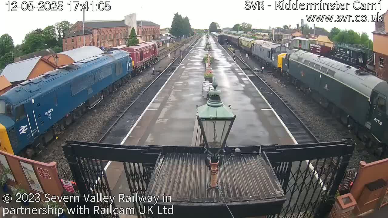 A wet railway station platform with multiple trains, some blue and some green, parked along the tracks.